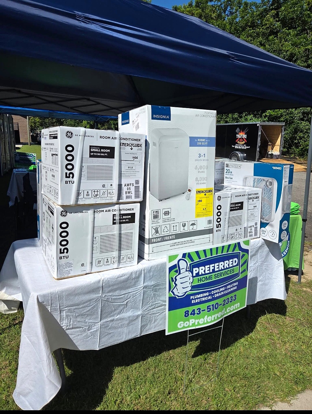 Boxed room air conditioners, a portable air conditioner, and box fans stacked on a draped table beneath an outdoor canopy.