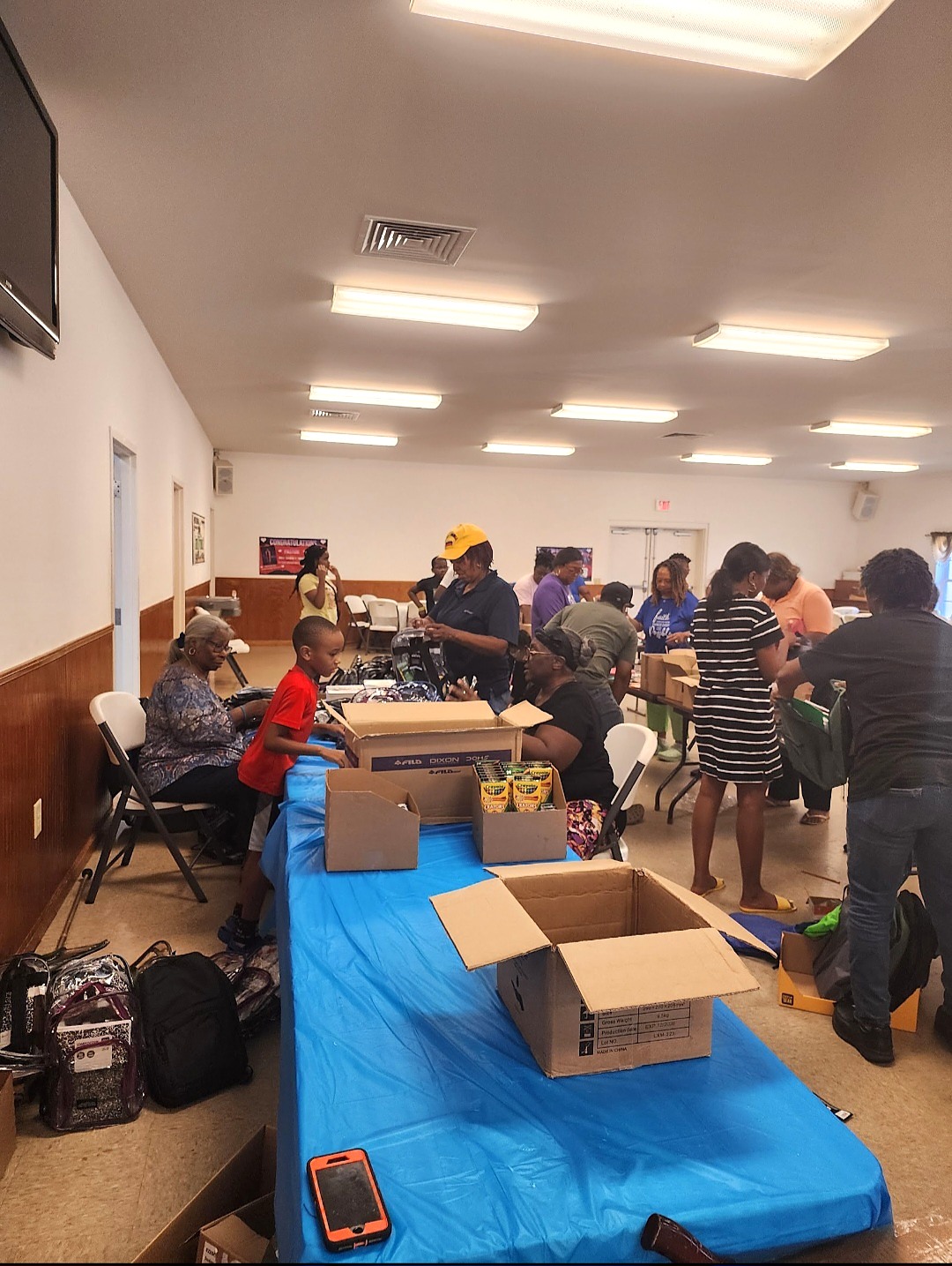 Volunteers of different ages sorting backpacks, crayons, and school supplies from cardboard boxes across long tables in a community hall.