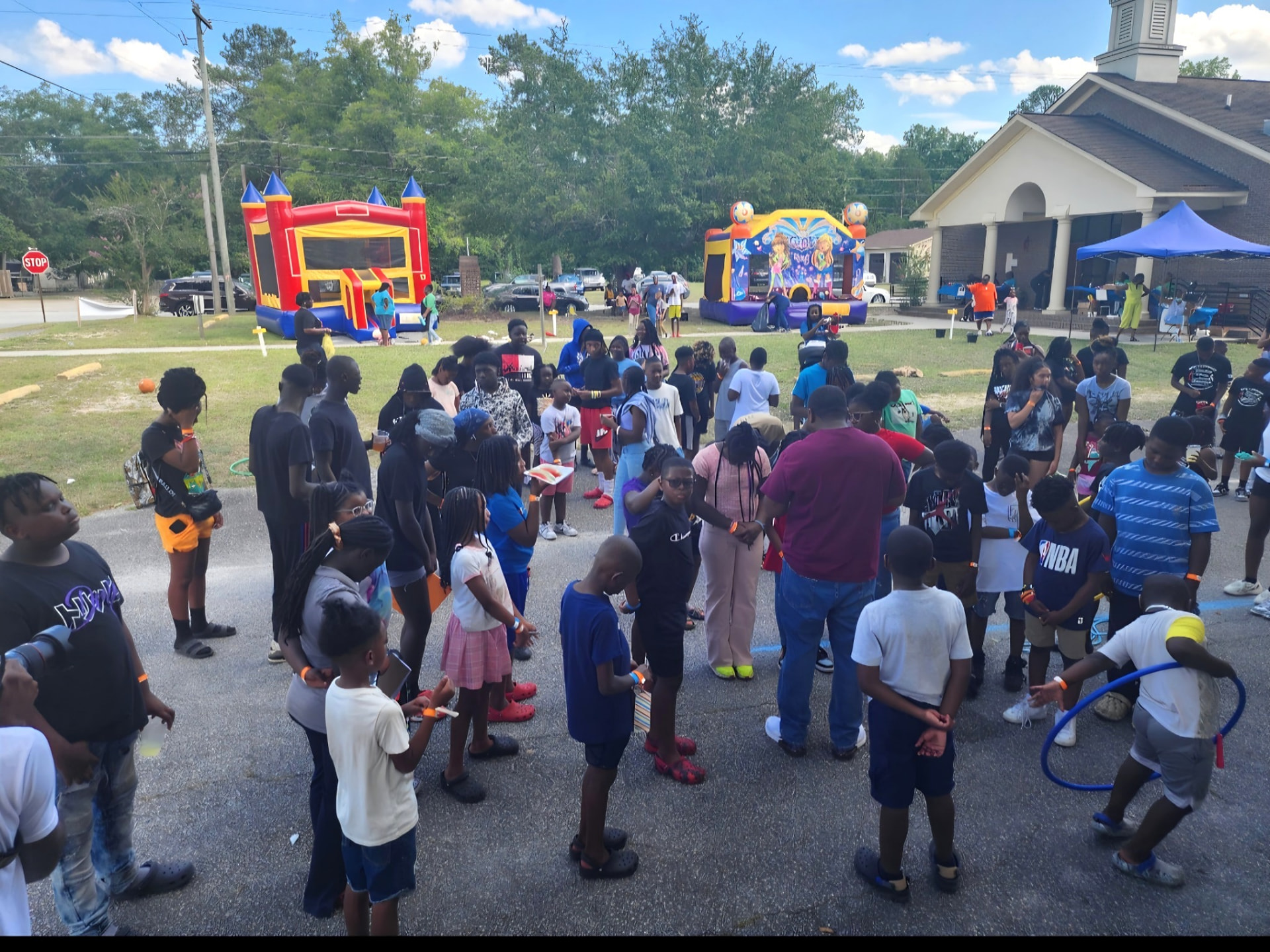 A large crowd of children and adults gathered on a paved lot beside inflatable bounce houses, a church building, and event tents.