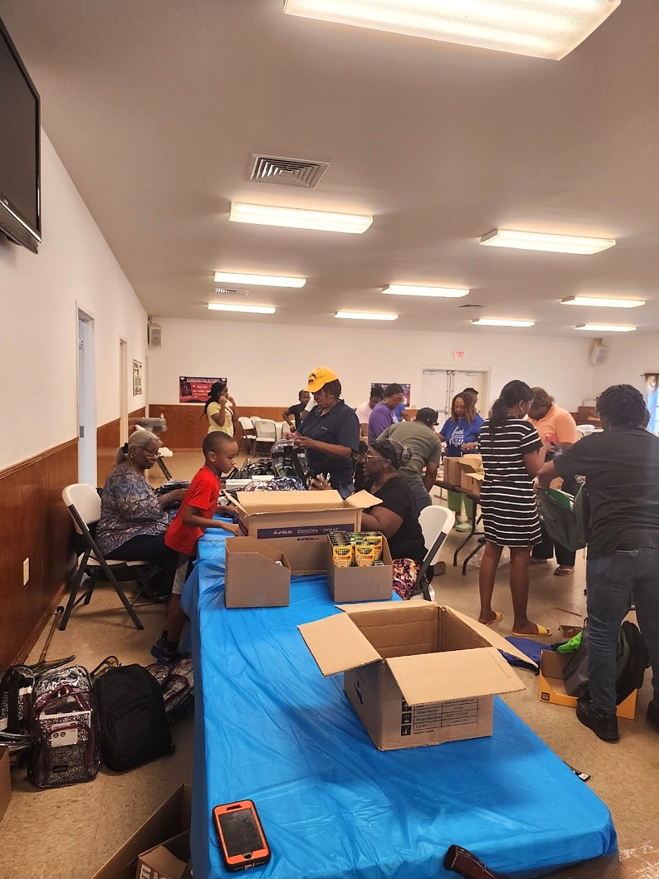 A wider view of volunteers filling backpacks with school supplies at tables inside a community hall.