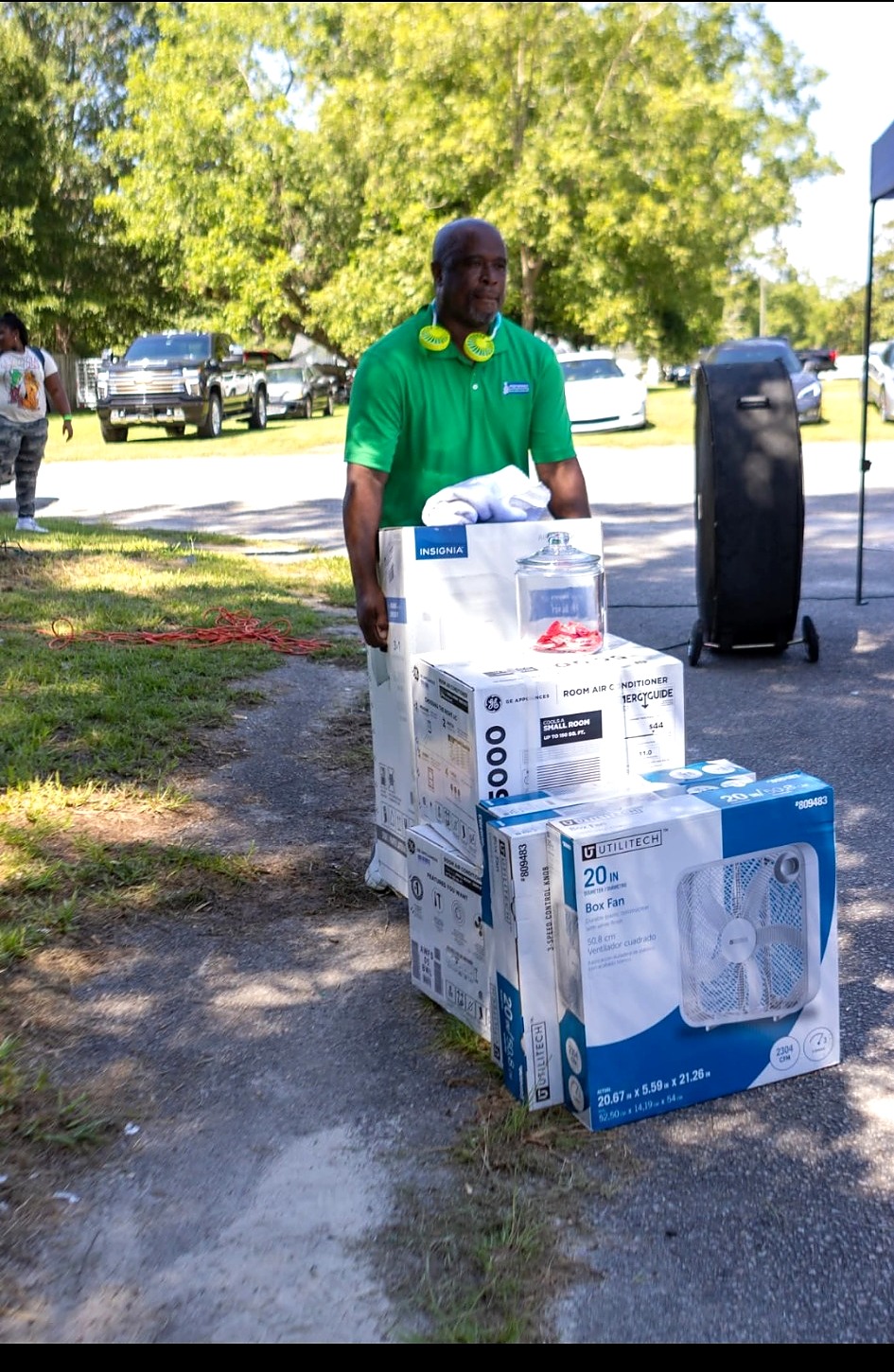 A man in a green polo standing outdoors beside a stack of boxed air conditioners and box fans ready for distribution.