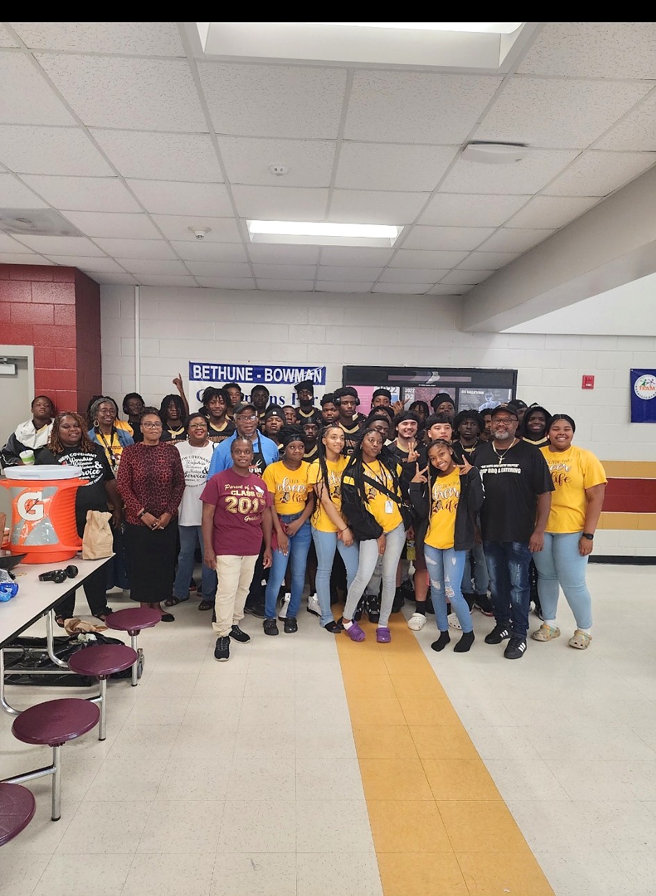 A group of students and adults posing together in a school cafeteria beneath a Bethune-Bowman banner.